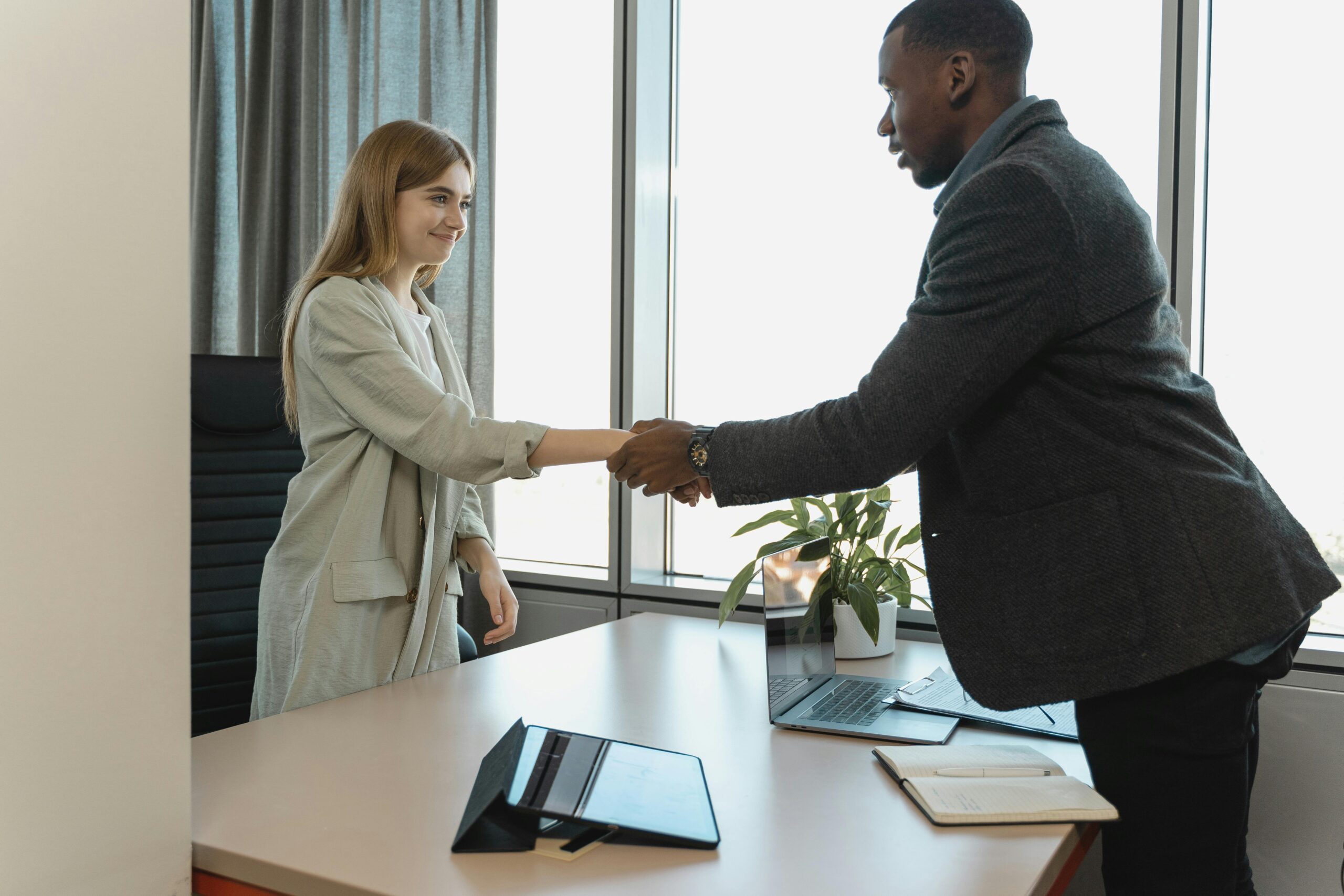 Two business professionals shaking hands across a desk in a bright office.