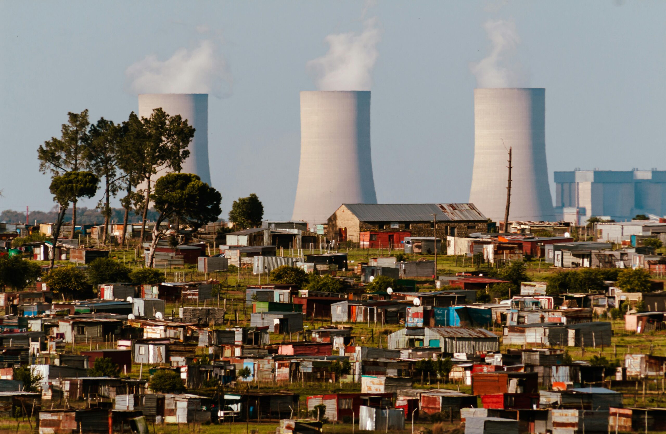 Cooling towers emit smoke over a vibrant South African settlement, highlighting industrial impact.