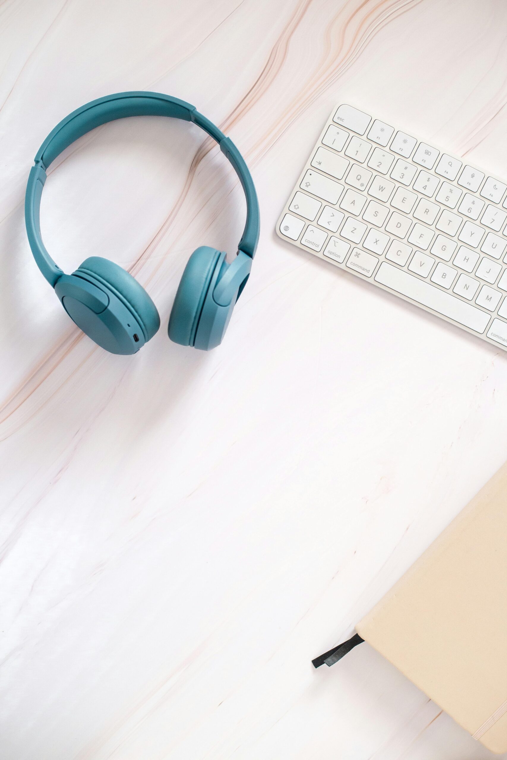 Flat lay of teal headphones, keyboard, and notebook on a desk.