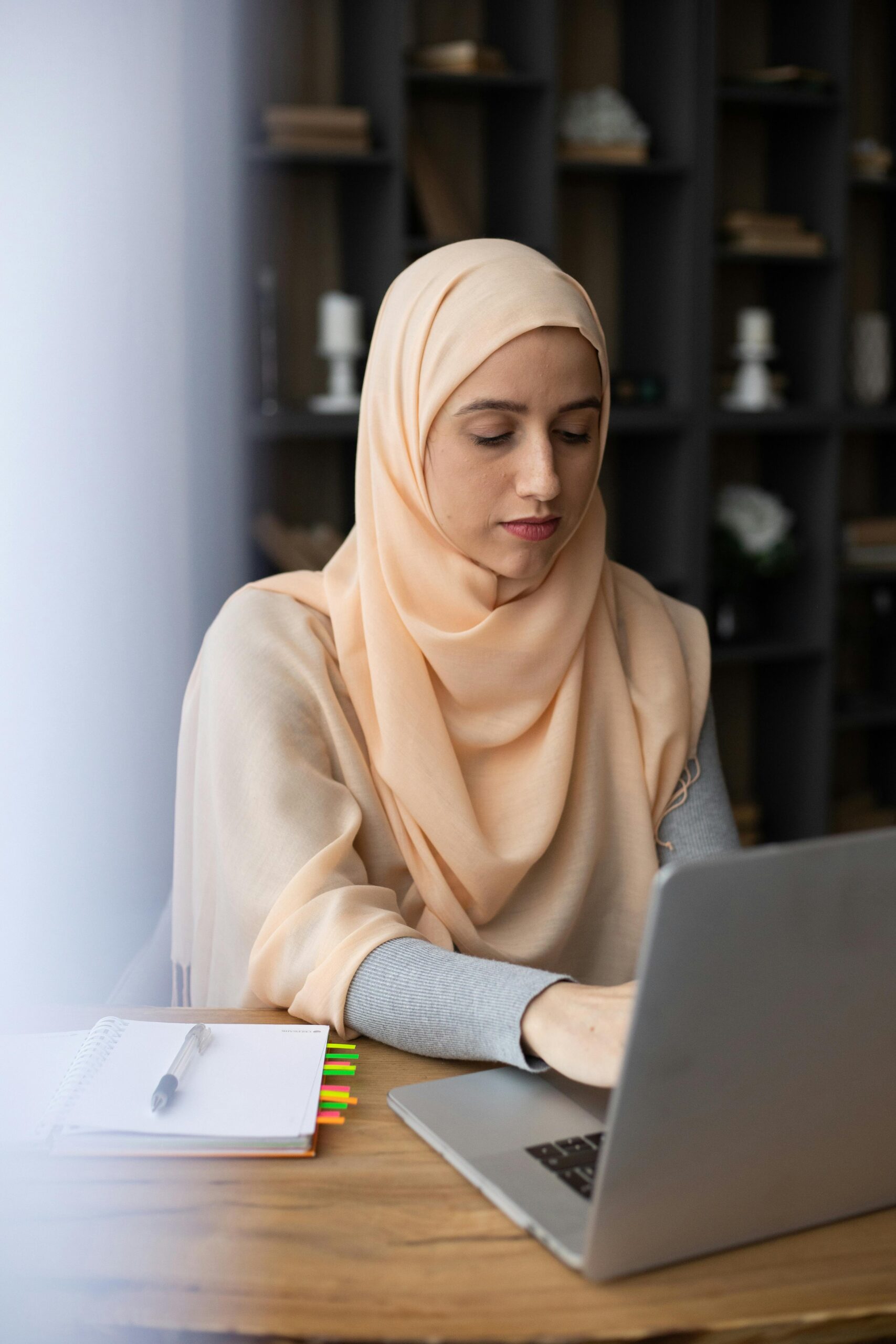 Muslim woman wearing a hijab works intently on her laptop indoors, surrounded by books.