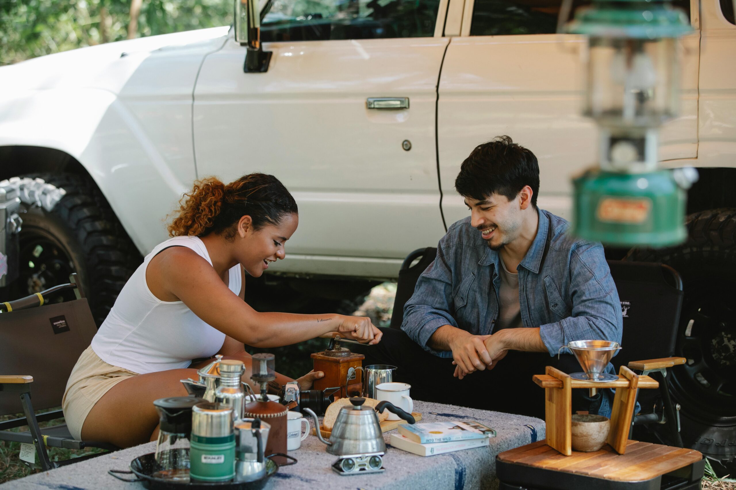 Cheerful young diverse couple in casual outfits grinding coffee and boiling water in gooseneck kettle while camping together in sunny nature