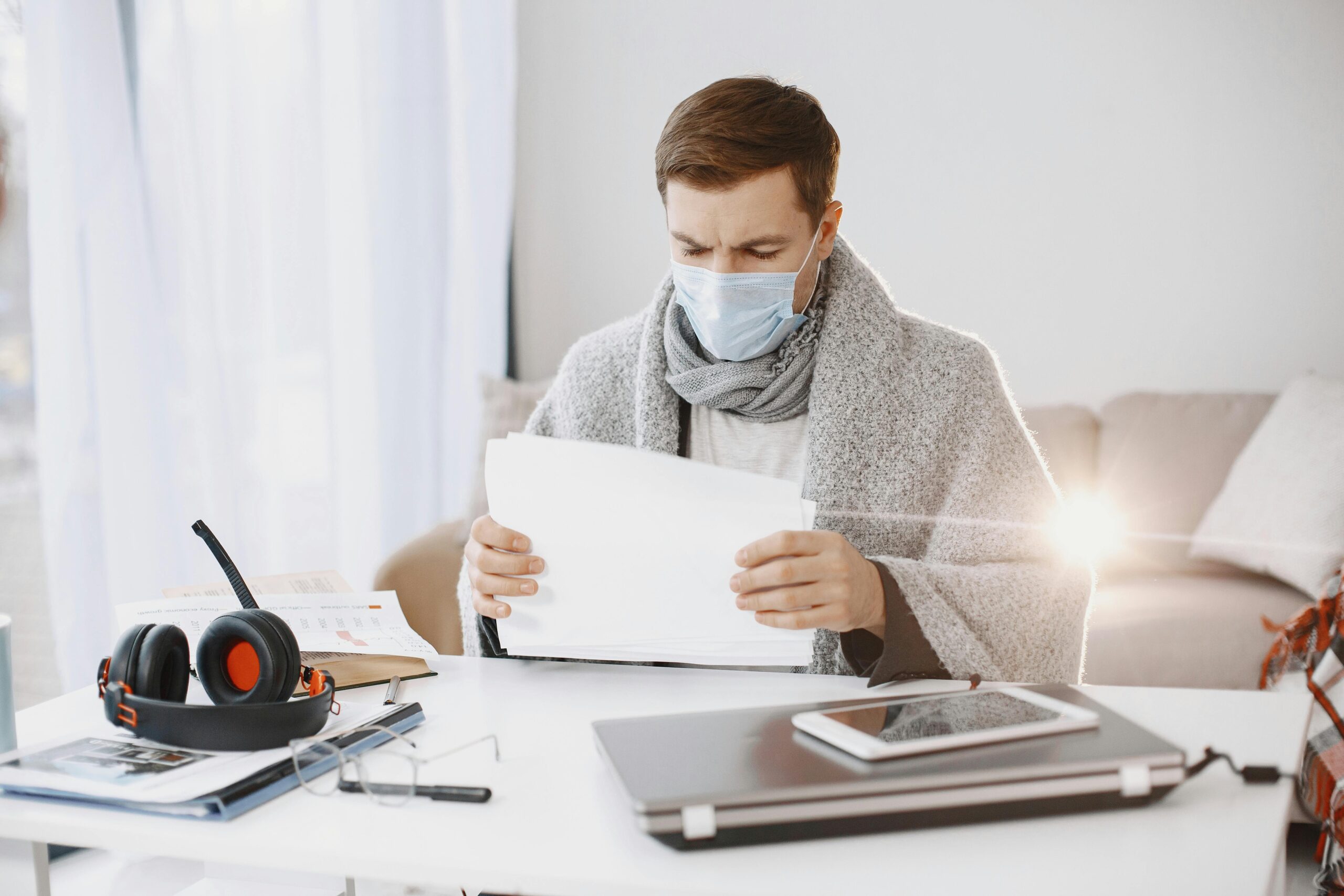 A man wearing a protective mask and scarf works from home, surrounded by office supplies on a bright day.