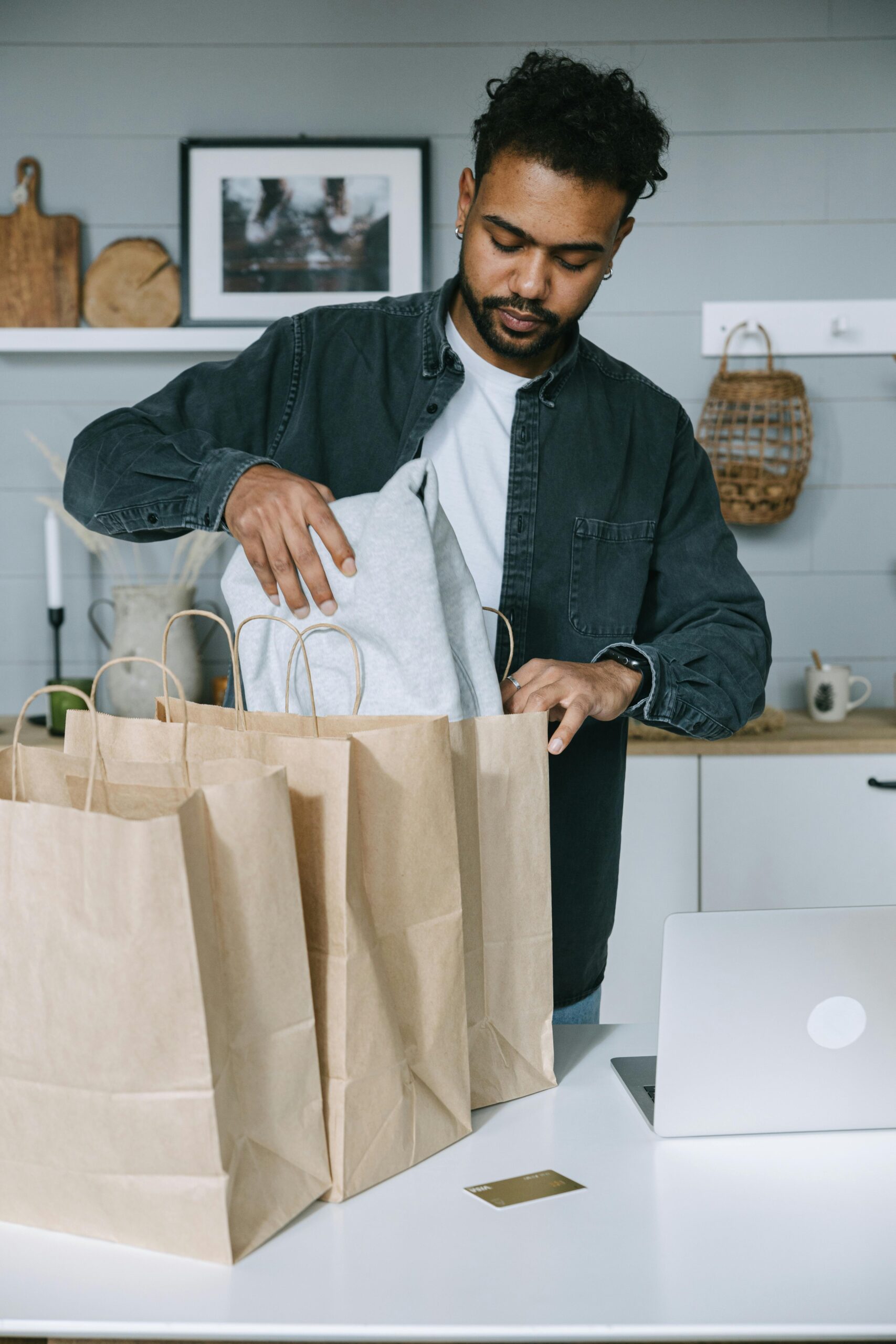 Adult man organizing purchases with paper bags and laptop in a modern home setting.