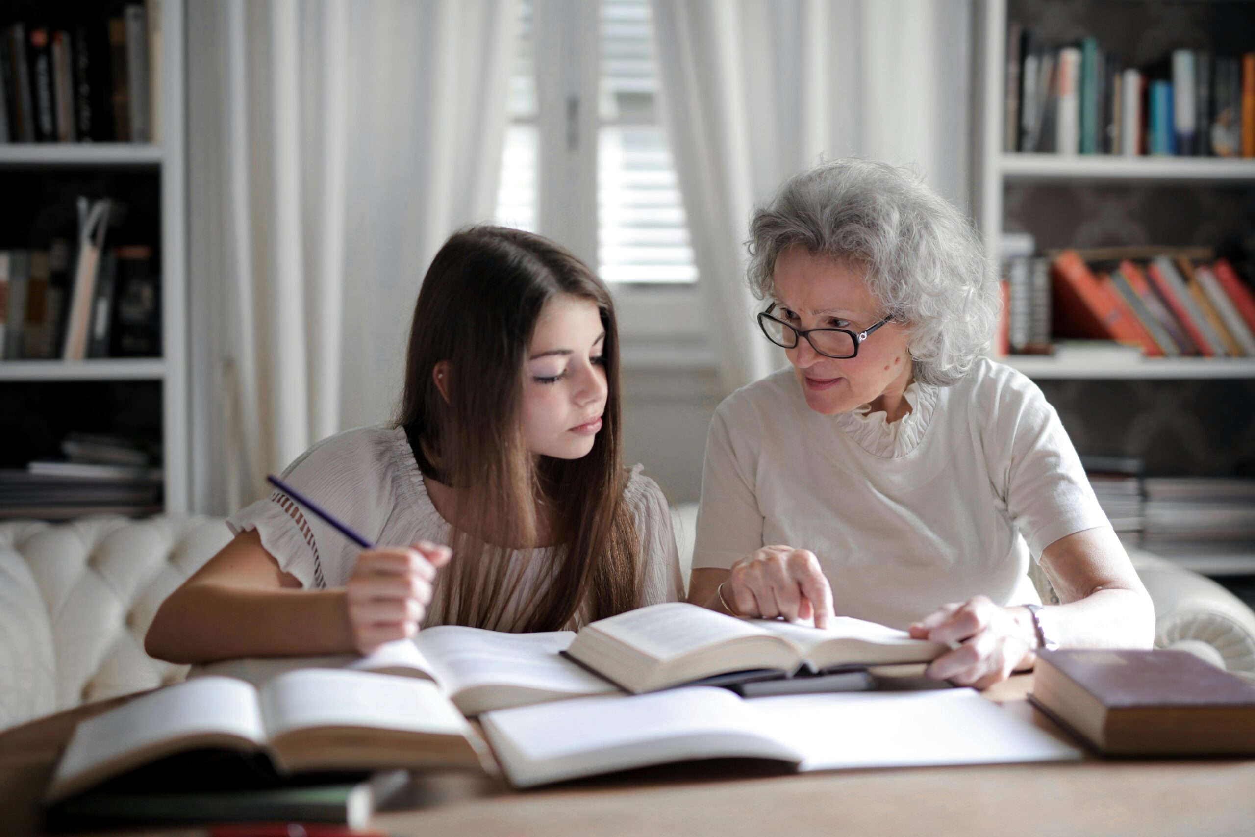 A grandmother helps her teenage granddaughter with homework, surrounded by books indoors.