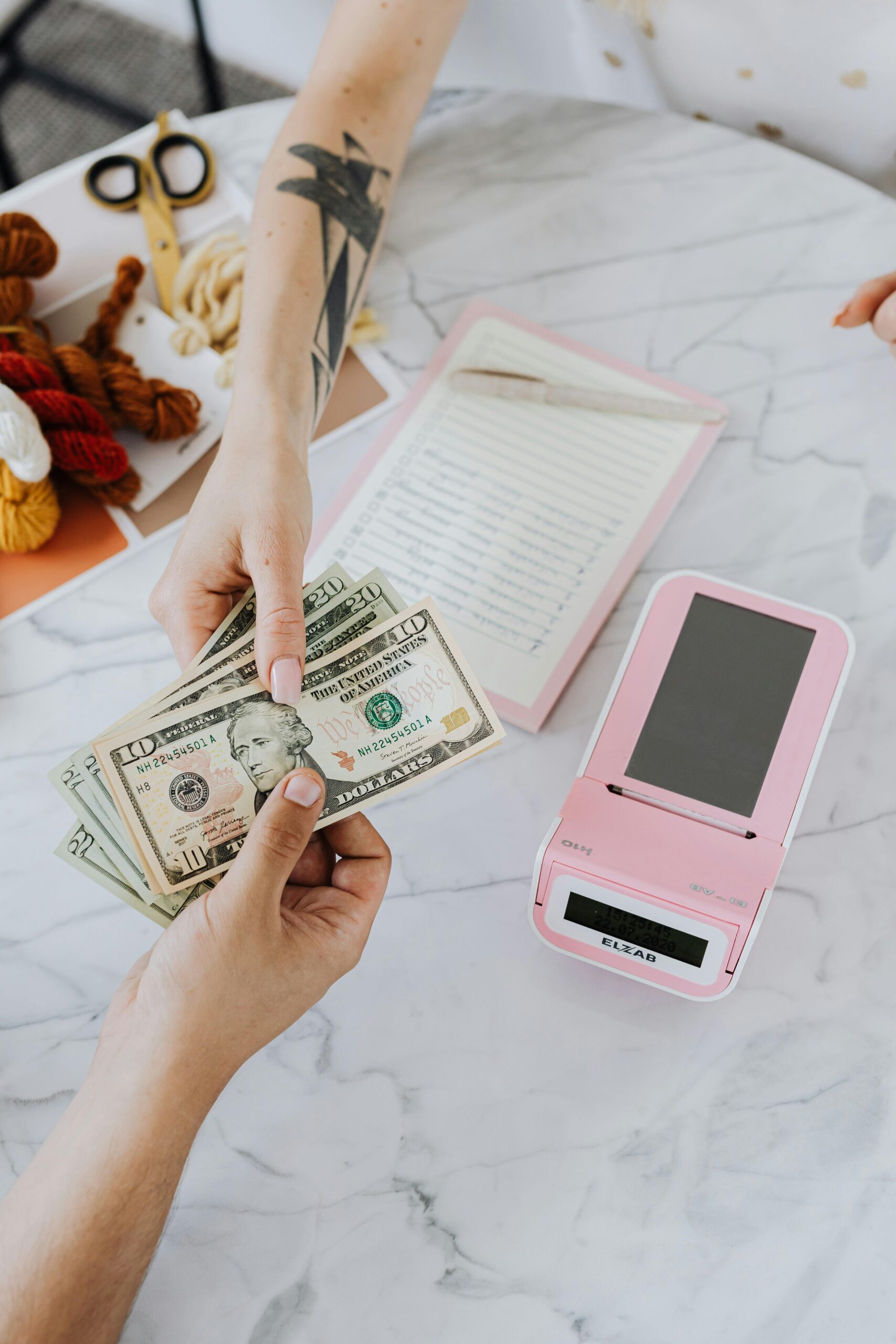 Hands exchanging US dollars with a pink card reader, notebook, and yarn on a marble table.