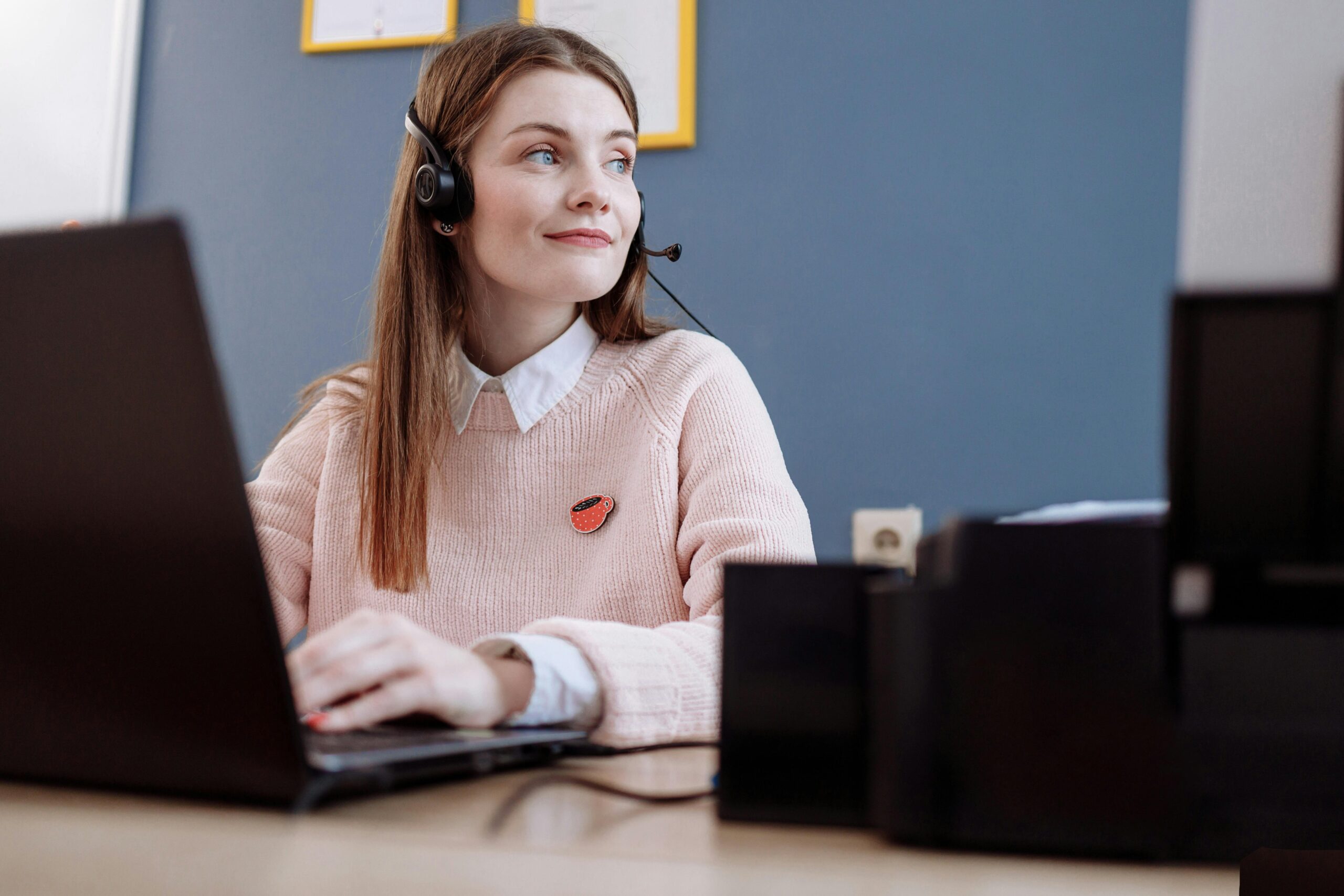 A cheerful woman in a pink sweater working remotely with a laptop and headset, providing customer service.