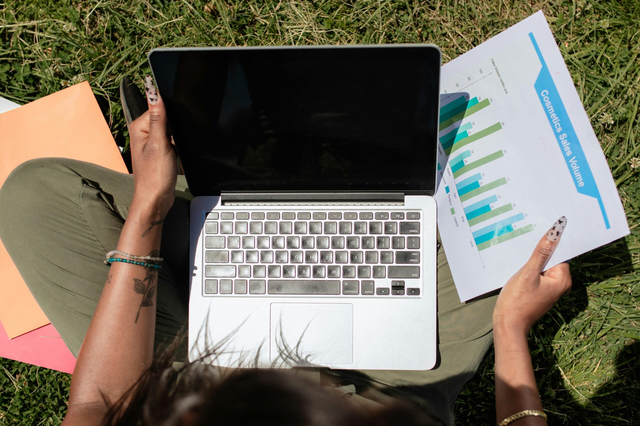 Woman reviewing sales charts on a laptop while sitting on grass, showcasing remote work flexibility.