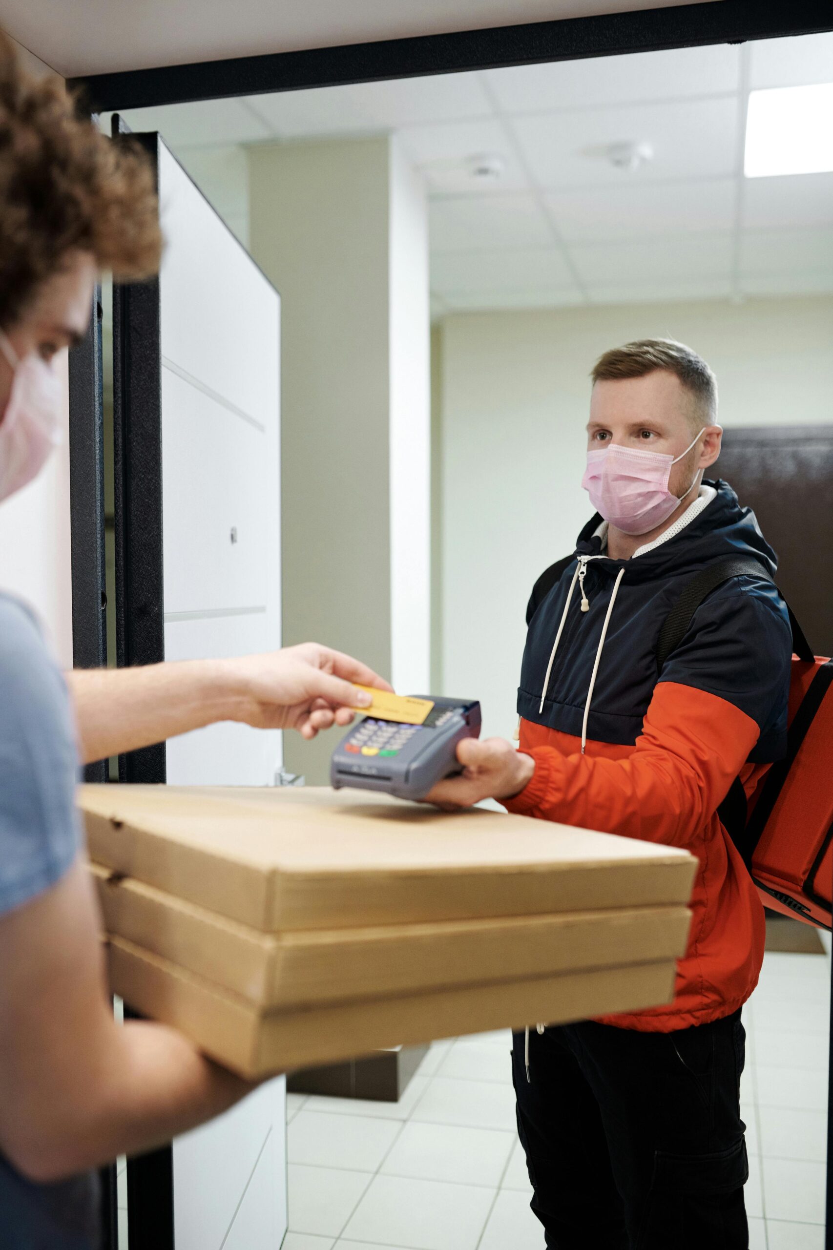 Deliveryman in mask handing pizza and credit card machine to a customer indoors.