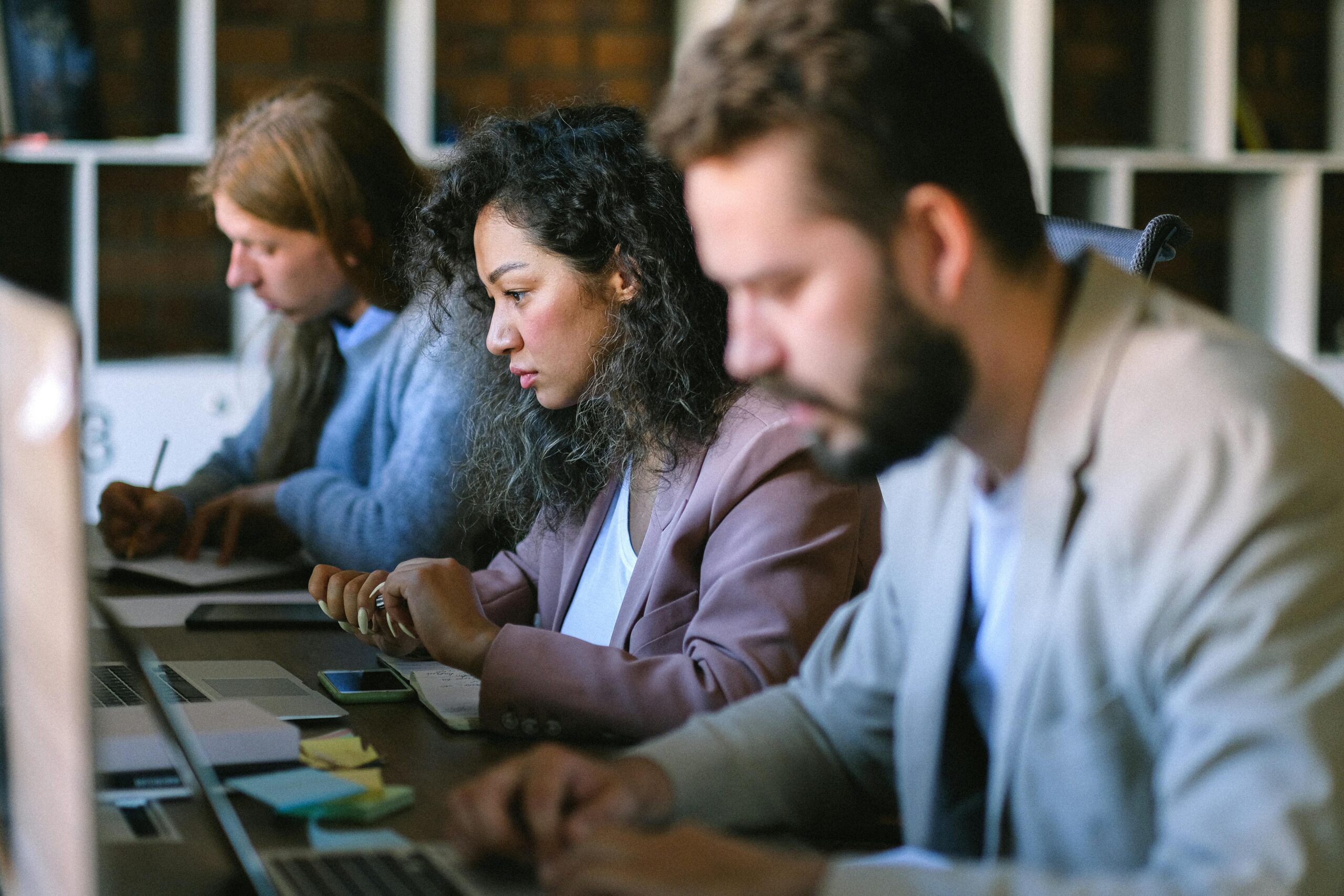 A diverse group of professionals collaborating in an open office space, using laptops and taking notes.